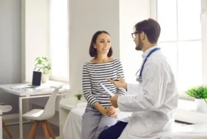 A doctor wearing a white coat and stethoscope talks to a woman sitting on an exam table in a bright medical office.