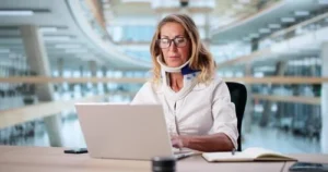 A woman wearing glasses and a neck brace works on a laptop at a desk with a notebook, inside a modern building with large windows.
