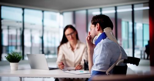 A man with a neck brace and arm sling sits at a desk talking to a woman with a laptop and clipboard in an office setting.