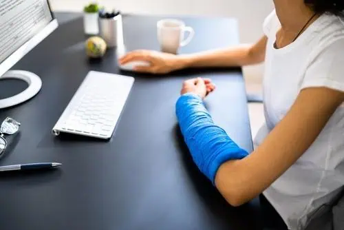 Person with a blue cast on their right arm sits at a desk using a computer keyboard, with a pen, glasses, and coffee mug nearby.