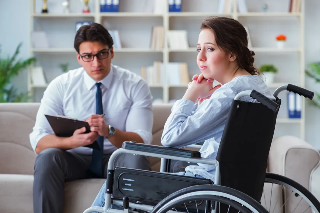 A woman in a wheelchair sits with her hands clasped, looking thoughtful, while a man in glasses with a clipboard sits across from her in an office setting.