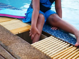 A person sitting poolside holds their lower leg, with one foot near an opening in the pool deck grating.