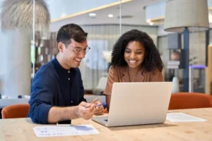 Two people sit at a table, smiling and looking at a laptop screen, with documents and a pen on the table in a modern office setting.