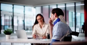 A man wearing a neck brace and arm sling sits at a desk speaking with a woman who is taking notes on a clipboard in an office setting.