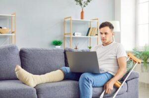Man with a bandaged leg and crutches sits on a gray sofa, using a laptop in a bright, modern living room.