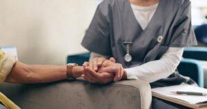 A healthcare worker in scrubs holds a patient's hand, offering comfort during a conversation. A clipboard and pen are visible nearby.