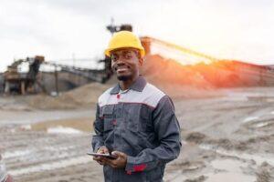 A man in a work uniform and yellow hard hat stands outdoors at an industrial site, holding a tablet and smiling at the camera.