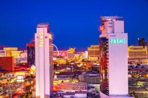 Two tall buildings labeled "PALMS" are lit up at night with the bright lights of the Las Vegas Strip and city skyline visible in the background.