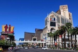 A sunny street view of the Las Vegas Strip featuring the Venetian, Palazzo, Wynn, and Treasure Island casino-hotels with palm trees and pedestrians.