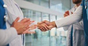 Two people in medical uniforms shake hands, while others in similar attire stand nearby and clap in a modern building.