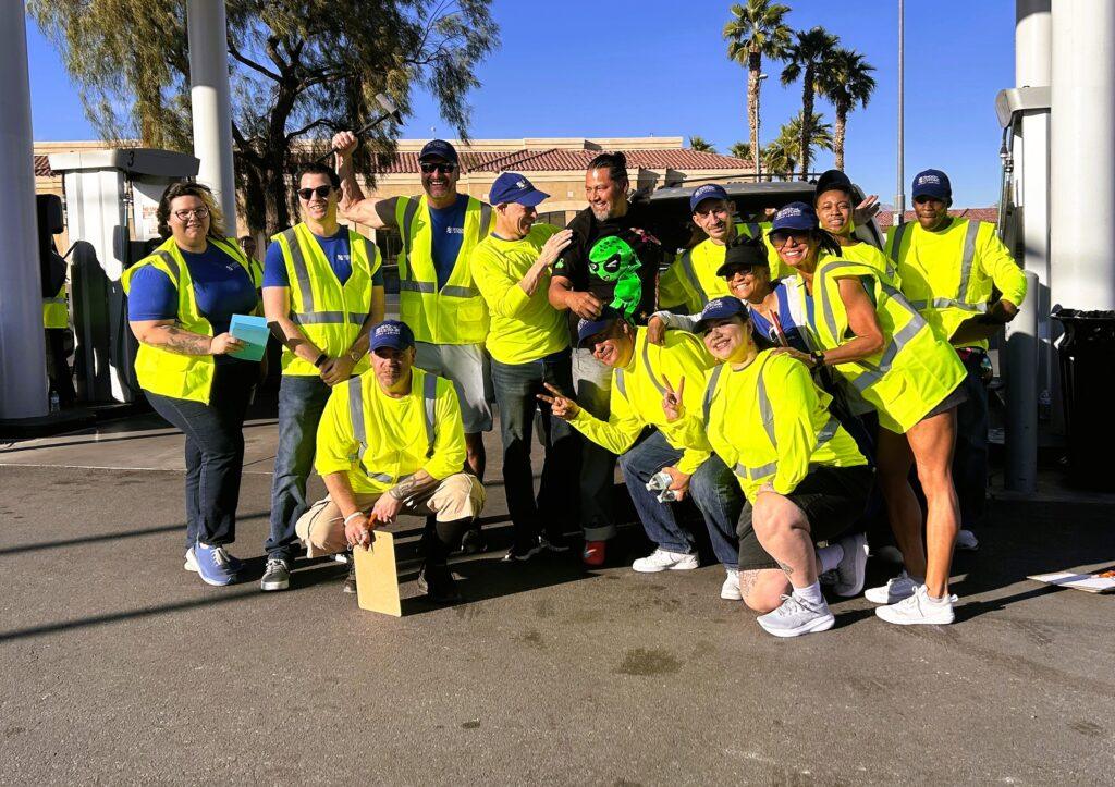 A group of people wearing yellow safety vests and blue hats pose together outdoors on a sunny day, some crouching and some standing, with palm trees in the background.