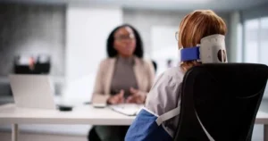 A woman wearing a neck brace and arm sling sits across from another woman at a desk in an office setting, having a conversation.