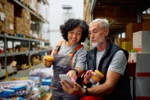 Two warehouse workers holding coffee cups look at a smartphone together, standing in an aisle surrounded by shelves filled with boxes and supplies.