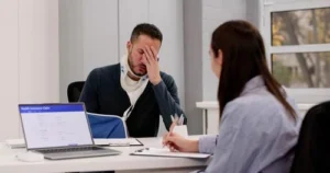 A man wearing a neck brace sits at a desk with his hand on his face, while a woman takes notes across from him. A laptop displaying health insurance data is on the desk.