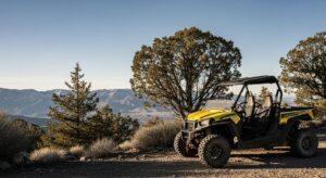 Yellow off-road vehicle parked on a dirt trail, surrounded by trees, with mountains visible in the background under a clear sky.