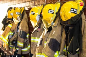 Five yellow firefighter helmets and turnout coats are hanging on hooks against a brick wall.
