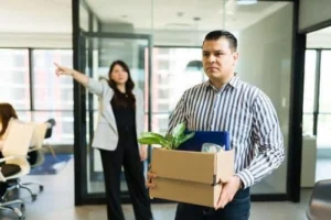 A man carrying a box of personal items leaves an office while a woman in the background points towards the exit.