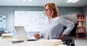 A woman in glasses sits at a desk with a laptop, holding her lower back and appearing to be in discomfort. Books and papers are on the desk, and a whiteboard is in the background.