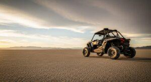 A black off-road UTV is parked on a vast, dry cracked desert landscape under a partly cloudy sky at sunset.