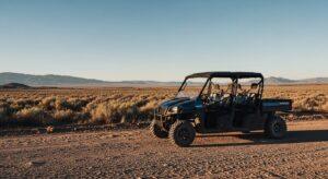 A black off-road utility vehicle is parked on a dirt road in a desert landscape with mountains visible in the background under a clear sky.