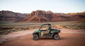 A green and black off-road vehicle is parked on sandy terrain with rocky desert cliffs and sparse vegetation in the background.