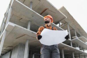 Man wearing an orange hard hat and gloves holding a blueprint, standing in front of a partially constructed concrete building.