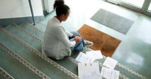 A person sits on indoor stairs next to scattered papers and an open binder, appearing deep in thought.