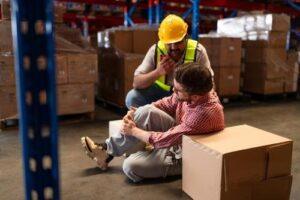 A worker sits on the warehouse floor holding his knee in pain while another worker in a safety vest and helmet calls for help.