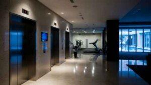A modern hallway with three closed elevator doors on the left and a gym with exercise equipment visible through glass doors on the right.