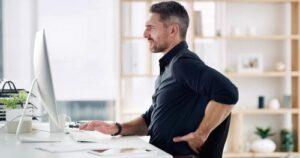 Office worker seated at computer, clutching lower back beside desk keyboard