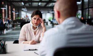 A woman wearing a neck brace sits across from a man at a desk in an office, looking concerned and holding her forehead.