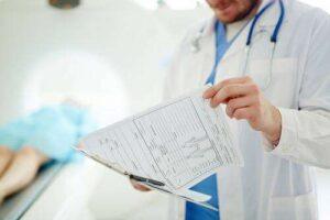 A doctor in a white coat reviews a patient’s medical chart on a clipboard, with a patient lying on a bed in the background.