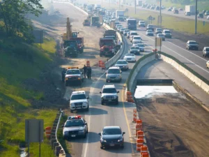 Highway under construction with traffic congestion, several police vehicles, construction equipment, and workers present; cars are merging due to lane closures marked with orange barrels.