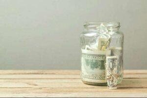 A glass jar filled with U.S. dollar bills sits on a wooden surface next to a rolled bundle of cash tied with string.