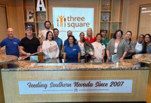 A group of people pose behind a kitchen counter at Three Square food bank, with a sign reading "Feeding Southern Nevada Since 2007" in front of them.