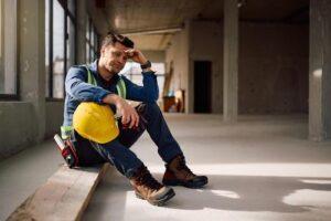distressed construction worker sitting on floor holding hard hat at job site